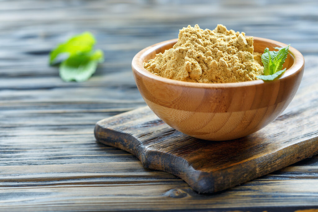 Wooden bowl filled with ground mustard powder on rustic wooden board, natural spice ingredient with fresh green mint leaves in background