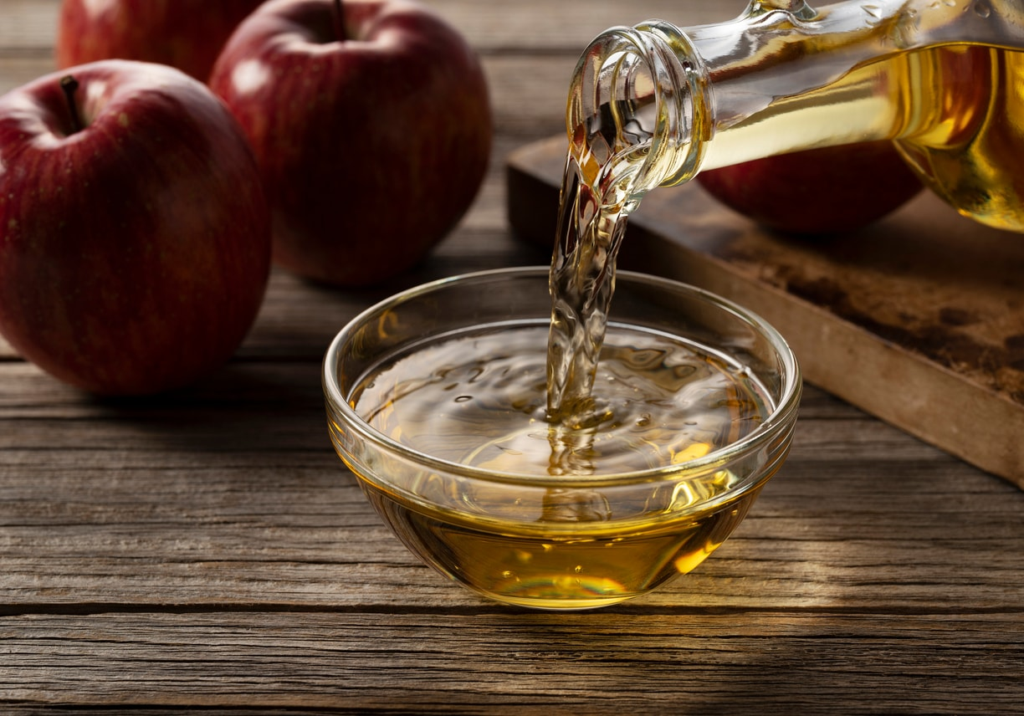 Apple cider vinegar being poured into a glass bowl with fresh red apples on rustic wooden table, natural health ingredient concept