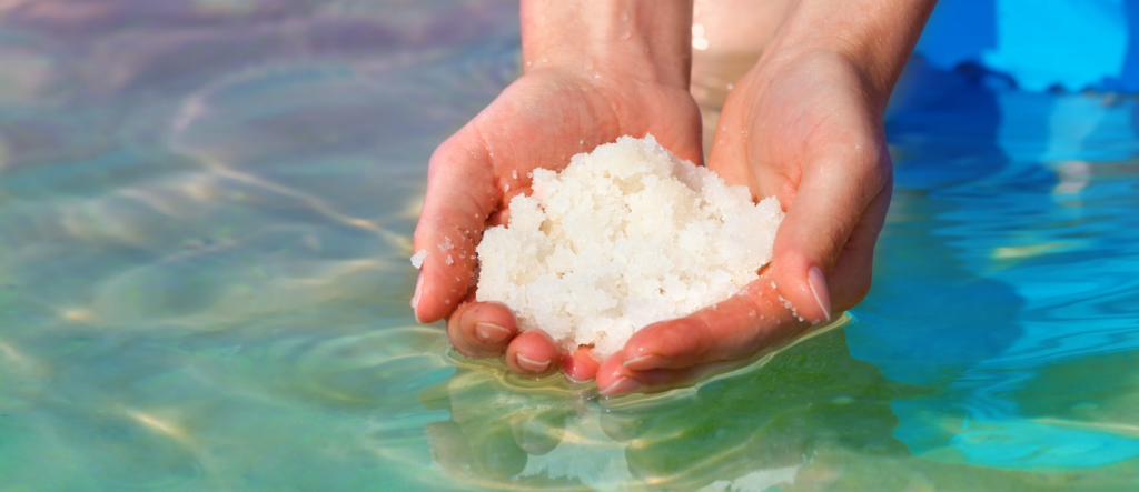 Hands holding Epsom salt crystals over clear blue bath water, natural magnesium sulfate spa and detox bath concept