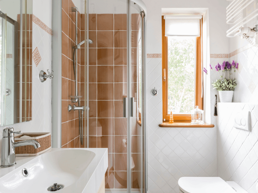 Compact bathroom with glass corner shower, warm terracotta tiles, white sink, toilet, and window bringing in natural light for a cozy, functional space.