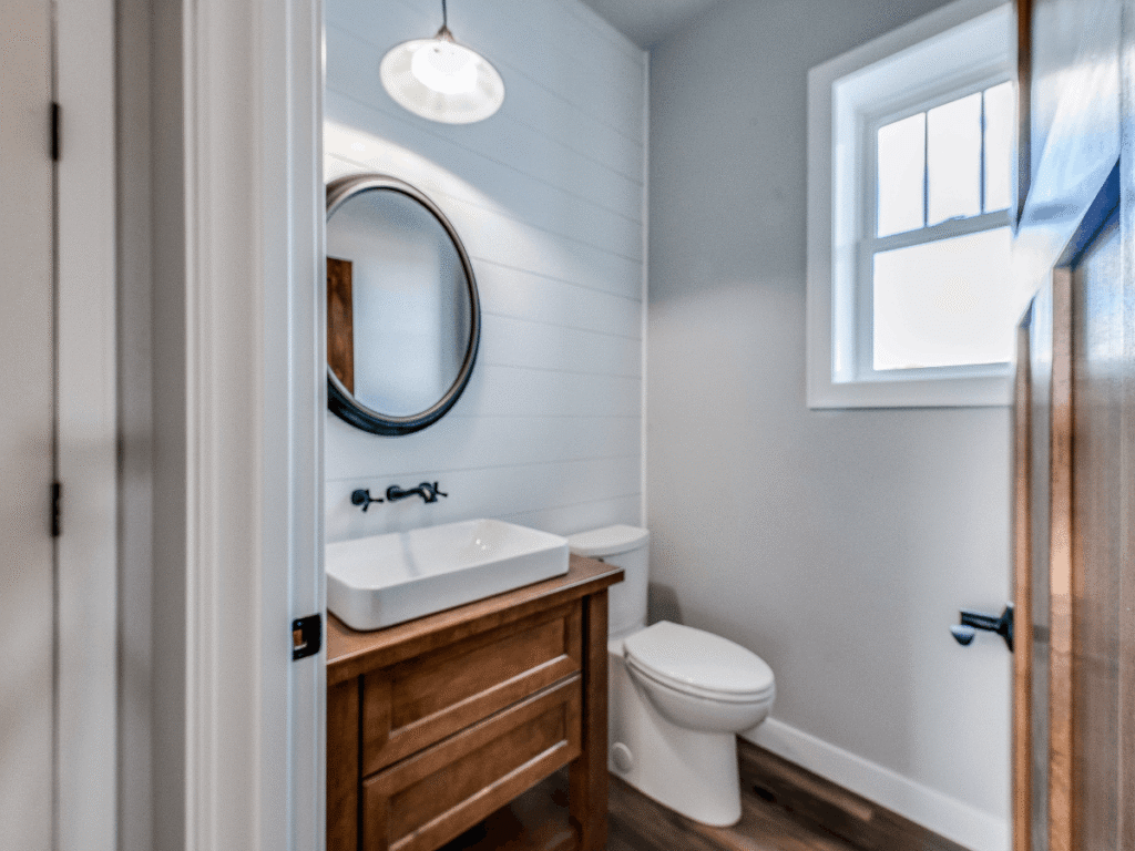 Compact bathroom with wood vanity, vessel sink, round mirror, white walls, and window bringing natural light into a clean, modern space.